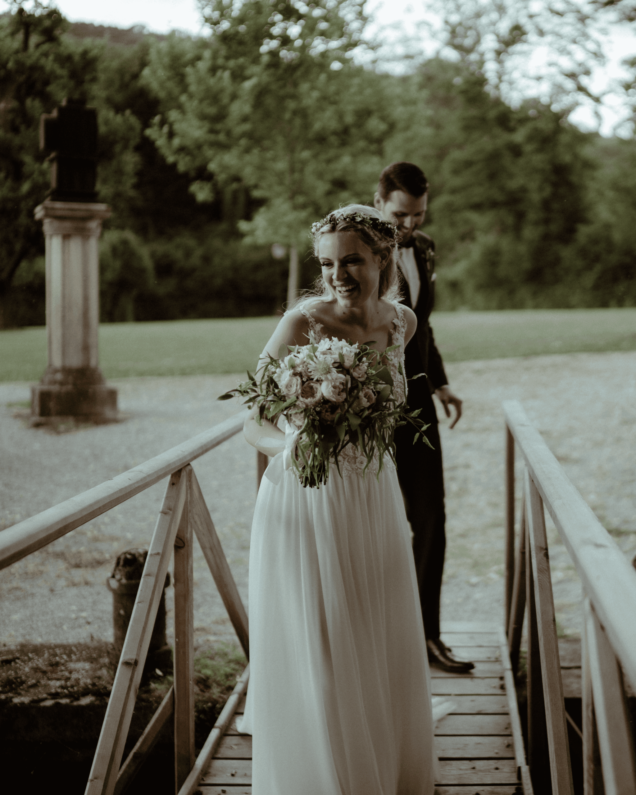 Bride walking across a wooden bridge, smiling, with the groom following in the background